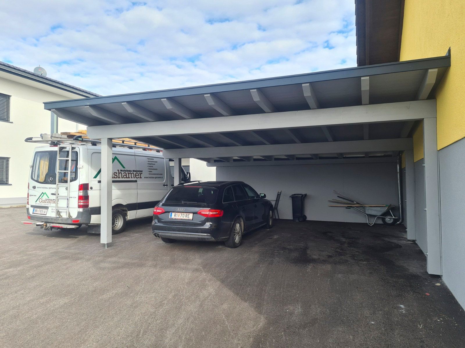 Ein Auto steht unter einem grauen Carport neben einem gelb-grauen Gebäude. Ein weißer Lieferwagen ist in der Nähe geparkt.