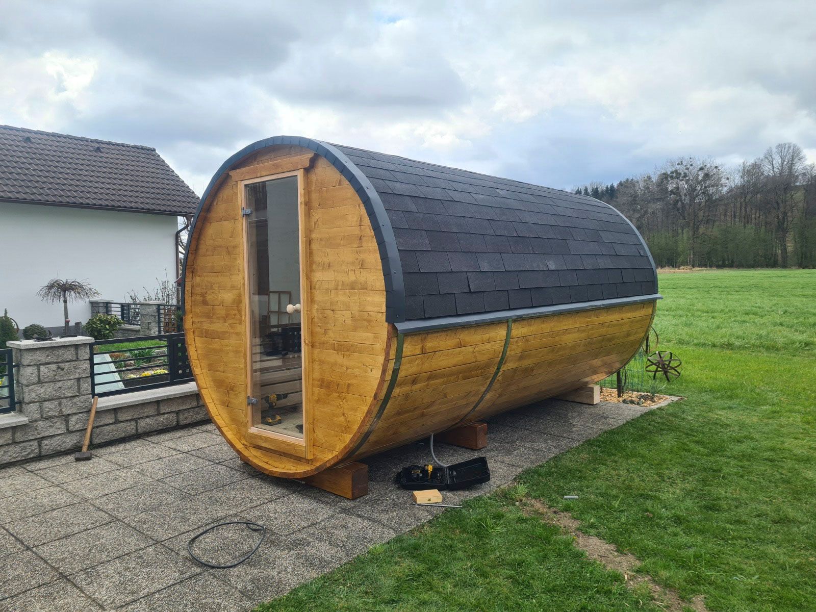 Eine hölzerne Fasssauna mit dunklem Schindeldach steht auf einer Beton-Terrasse in einem Hof.