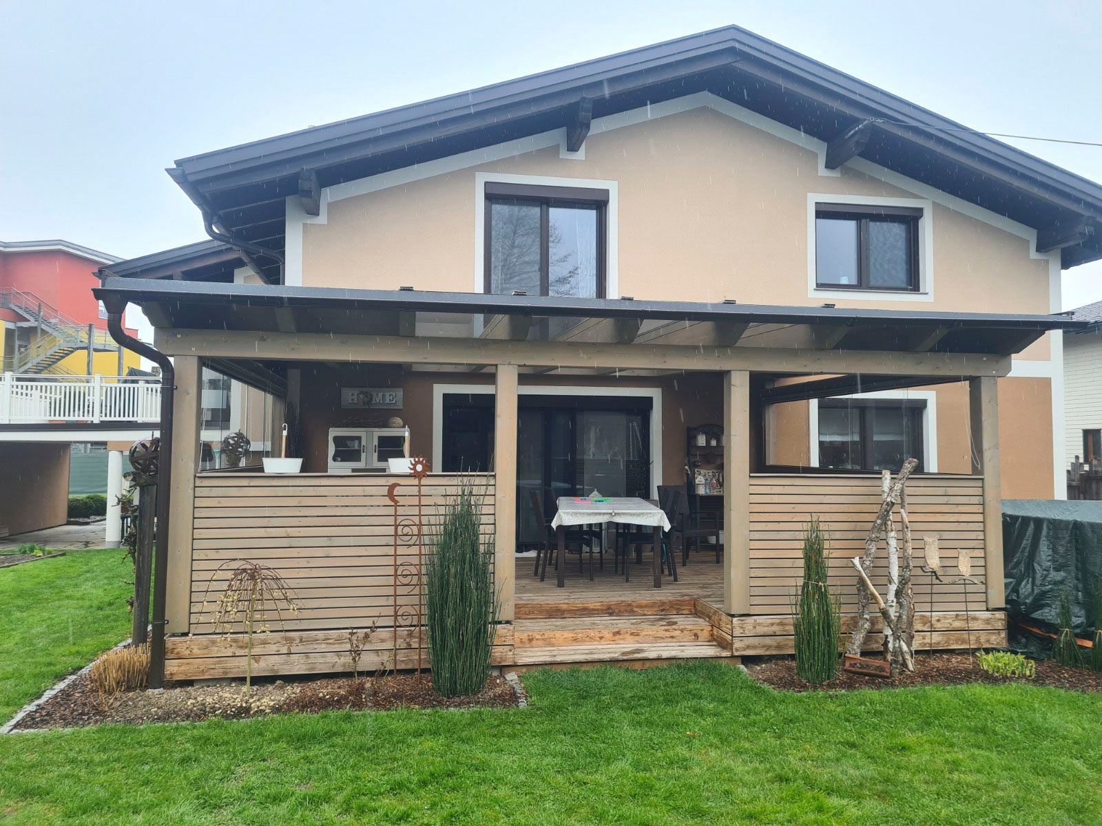 Beiges Haus mit überdachter Terrasse, Holzzaun und grünem Rasen.