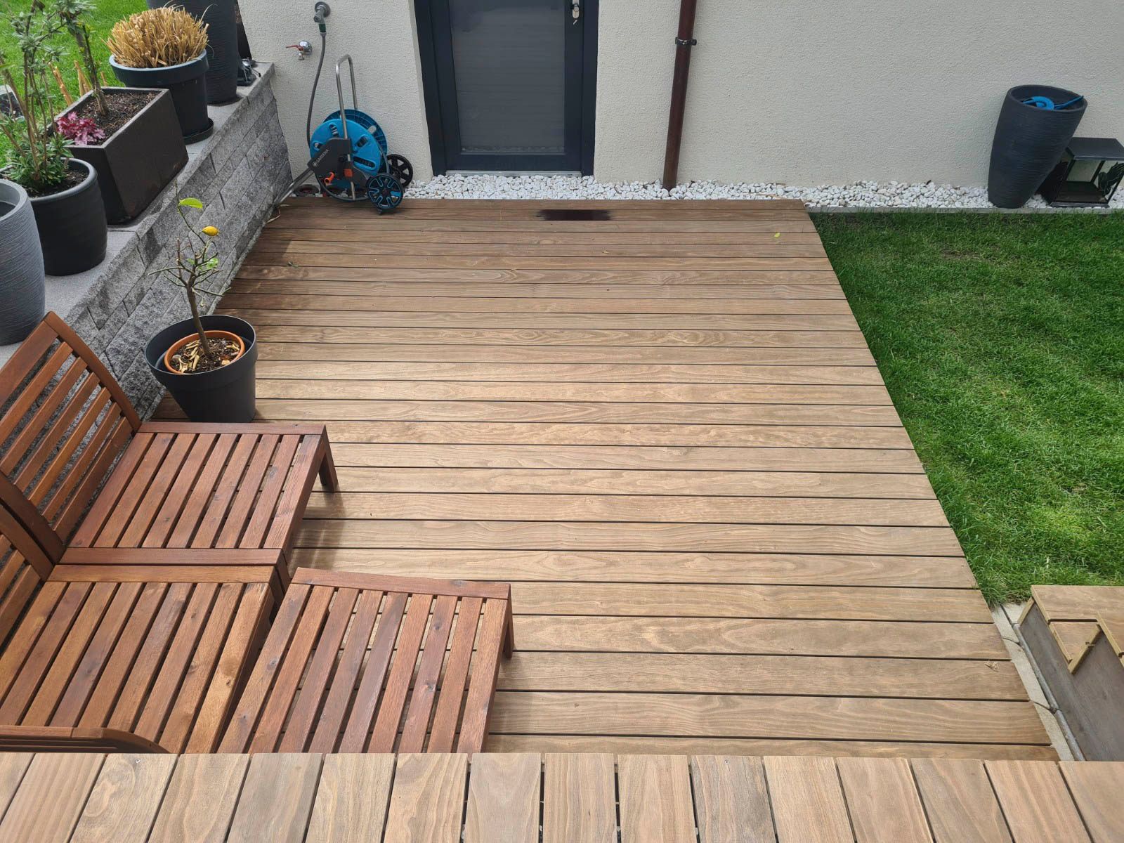 Holzterrasse mit Liegestuhl, neben einer Rasenfläche und einem Gebäude mit Topfpflanzen.