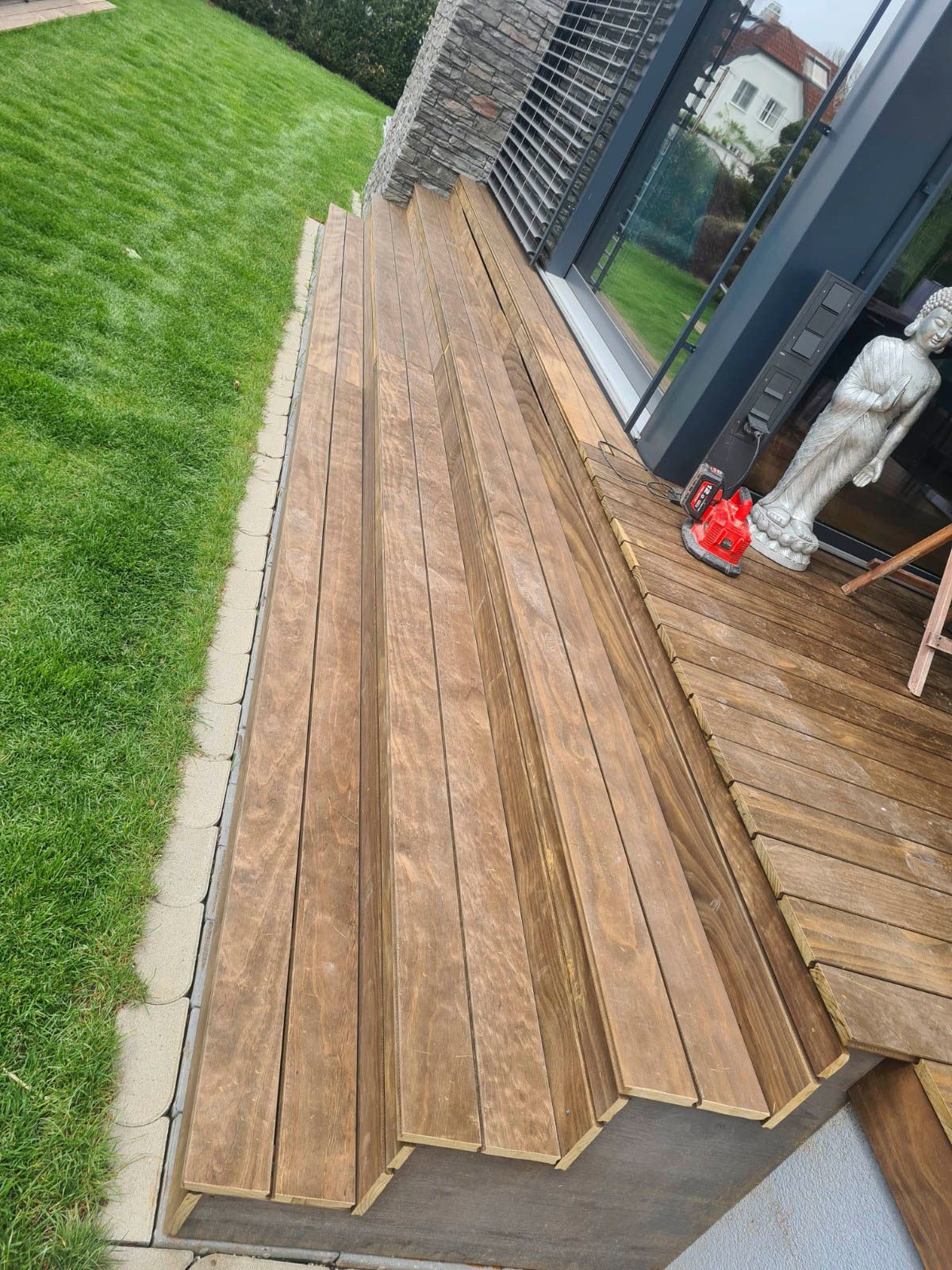 Holzterrasse mit Treppe neben einem Haus mit Statue und Rasenfläche.