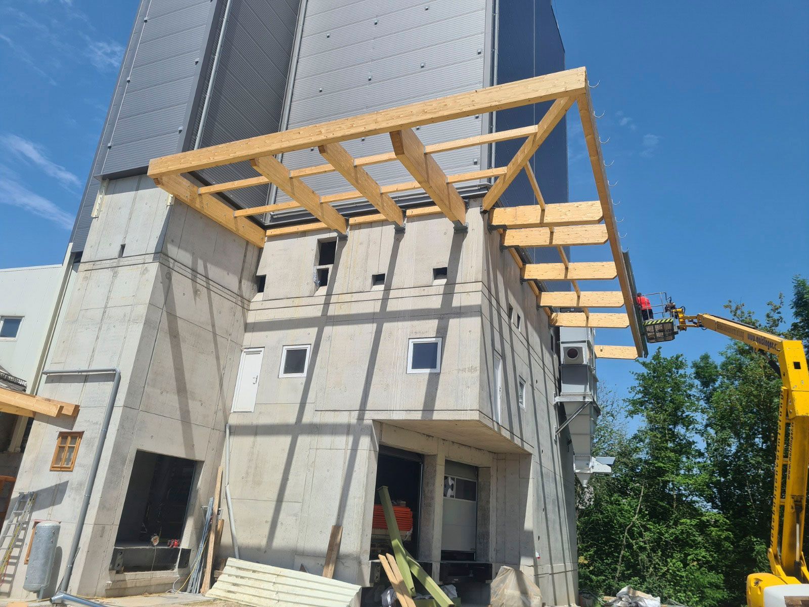 Errichtung eines hölzernen Vordachs auf einem Betongebäude, ein gelber Aufzug ist in Betrieb, strahlend sonniger Tag.