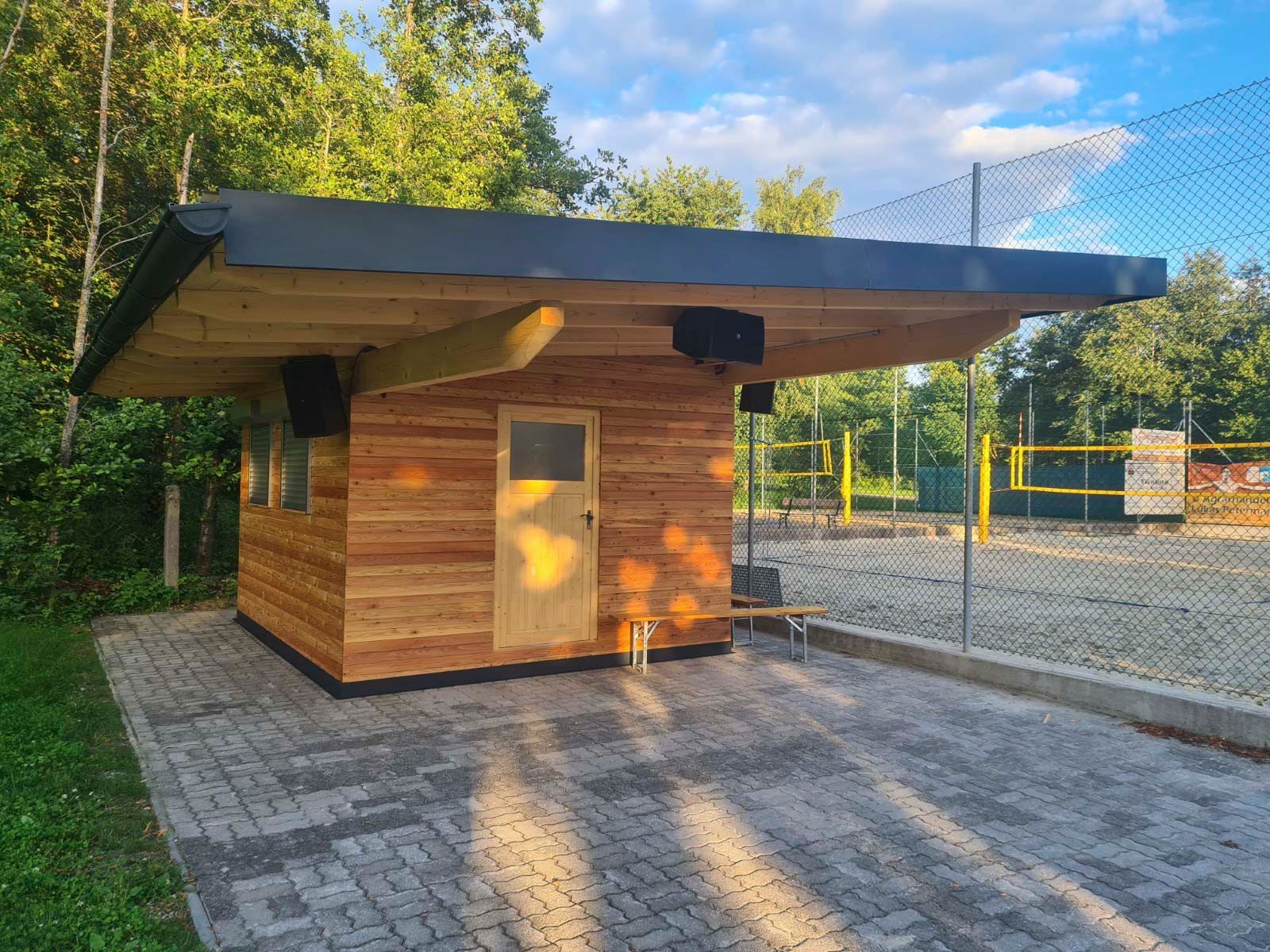 Holzgebäude mit schwarzem Dach, Tür, Fenstern und Tonanlage. Befindet sich neben einem Volleyballfeld.