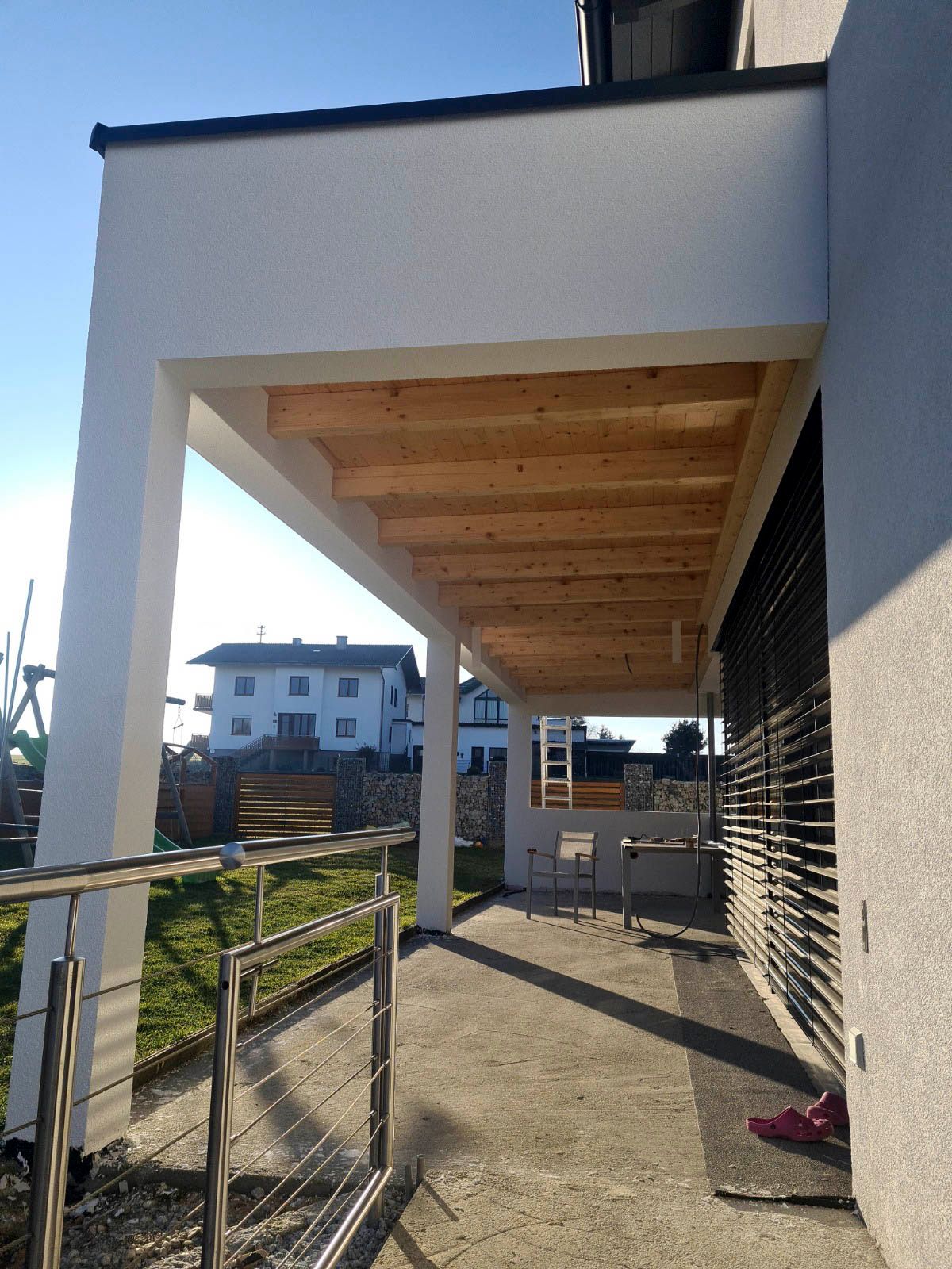 Überdachte Veranda mit weißen Säulen und Holzdecke, neben einem Haus mit einer Terrasse aus Beton.