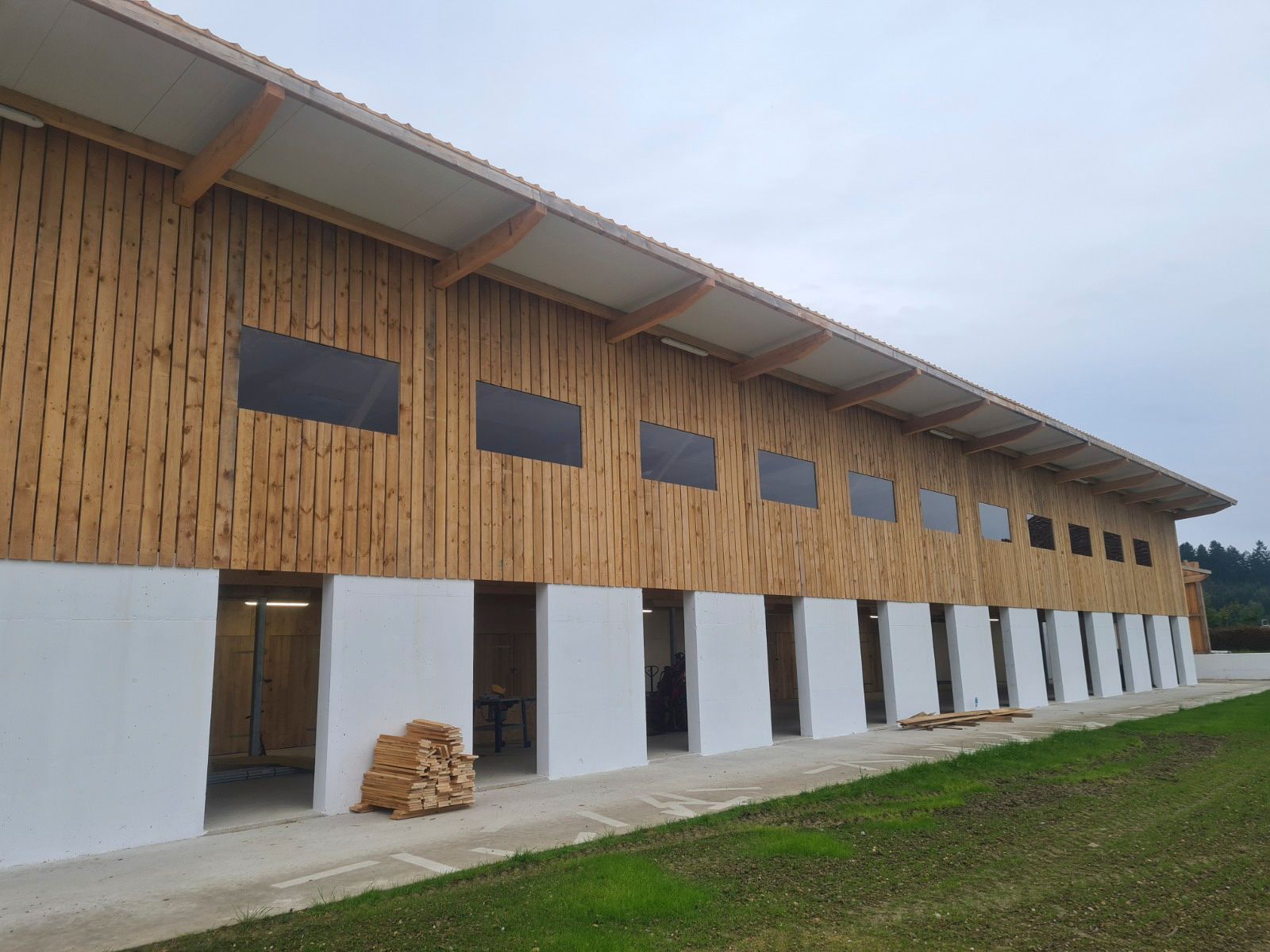 Lange Holzscheune mit weißen unteren Wänden und rechteckigen Fenstern; bedeckter Himmel.