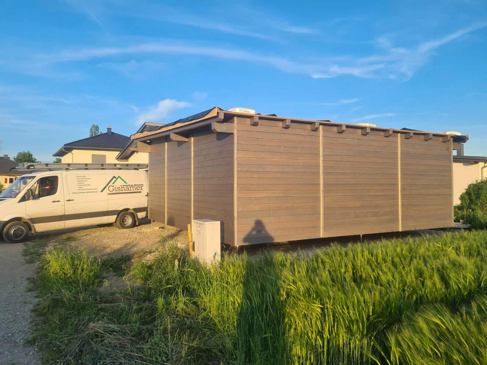 Braunes Gebäude mit Lieferwagen, Stromkasten und hohem Gras in sonniger Freiluftlage.