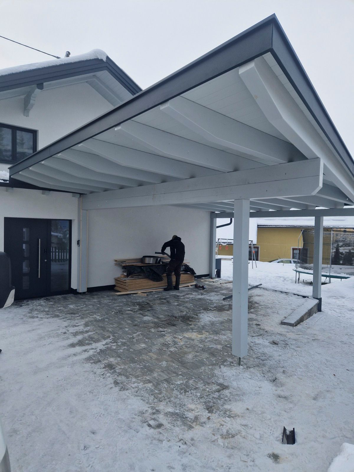 Eine Person arbeitet an einem im Bau befindlichen Carport neben einem schneebedeckten weißen Haus.