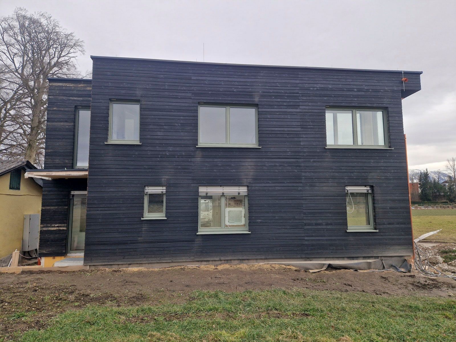 Schwarzes Holzhaus mit mehreren Fenstern. Bauschutt und Gras im Vordergrund. Bewölkter Himmel.