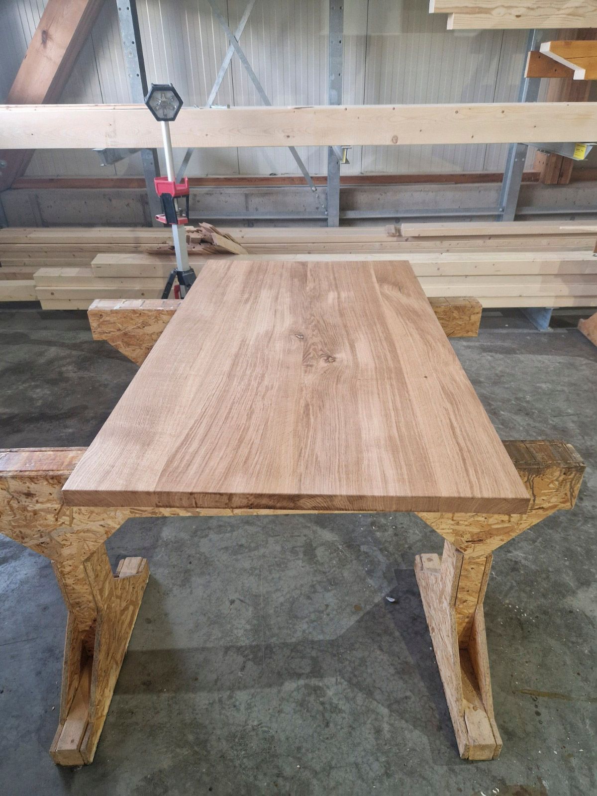 In einer Werkstatt ruht eine hölzerne Tischplatte auf zwei Sägeböcken. Im Hintergrund sind Bretter zu sehen.