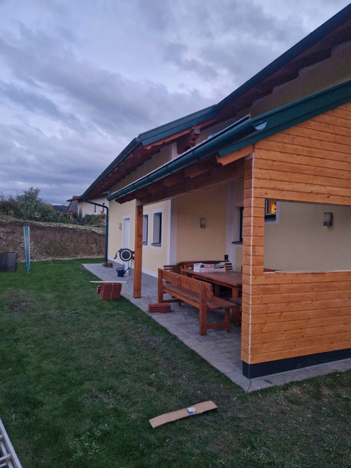 Hausfassade mit Holzverkleidung, überdachter Terrasse, Holztisch und -bank, grüner Rasen, bewölkter Himmel.