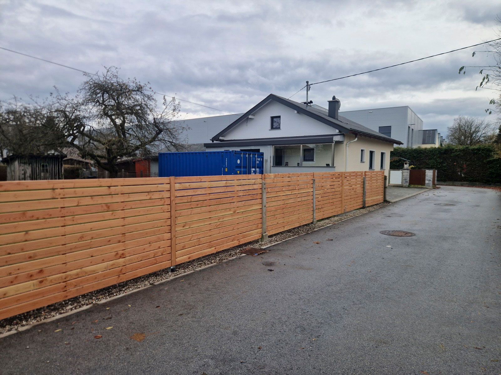 Ein helles Haus mit Holzzaun an einer Straße unter bewölktem Himmel.