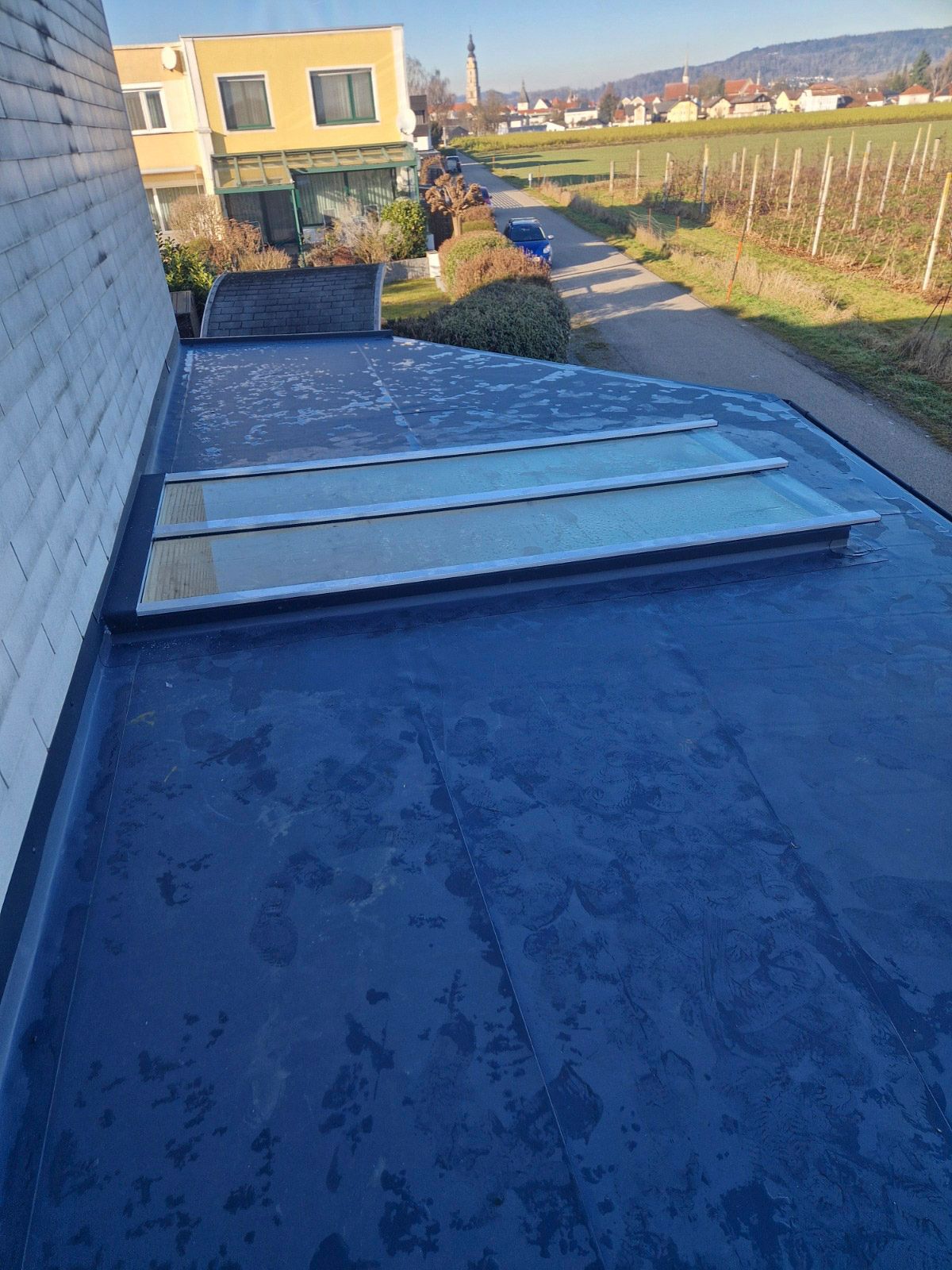 Flaches blaues Dach mit Oberlichtern, neben einer Backsteinmauer. Ein Weg führt zu einem Feld im Hintergrund.