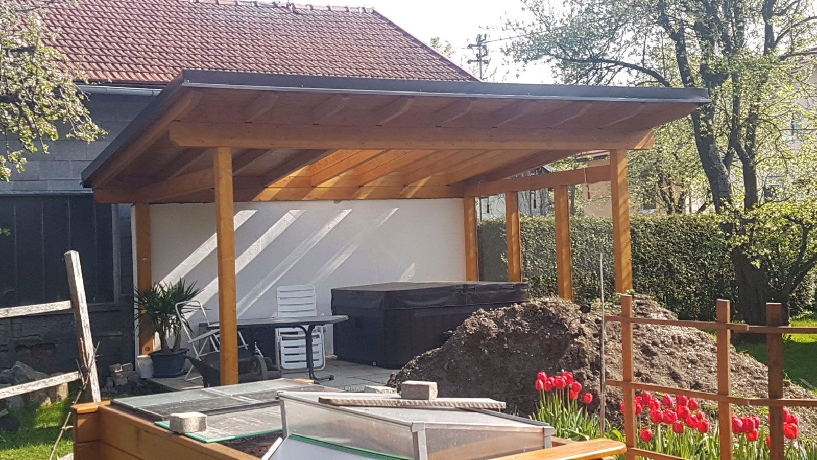 Eine hölzerne Pergola, die an ein Gebäude angebaut ist, mit einer weißen Innenwand und einem Erdhaufen.