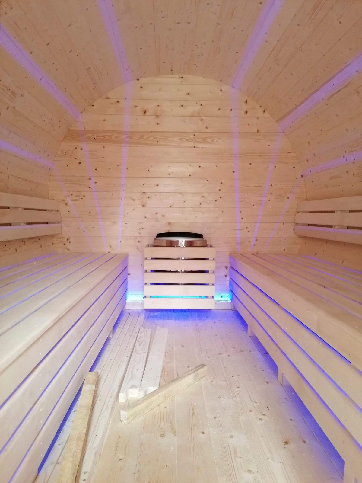 Innenansicht einer Sauna mit hellen Holzbänken, Wänden und einem Ofen, beleuchtet von blauen und violetten Lichtern.