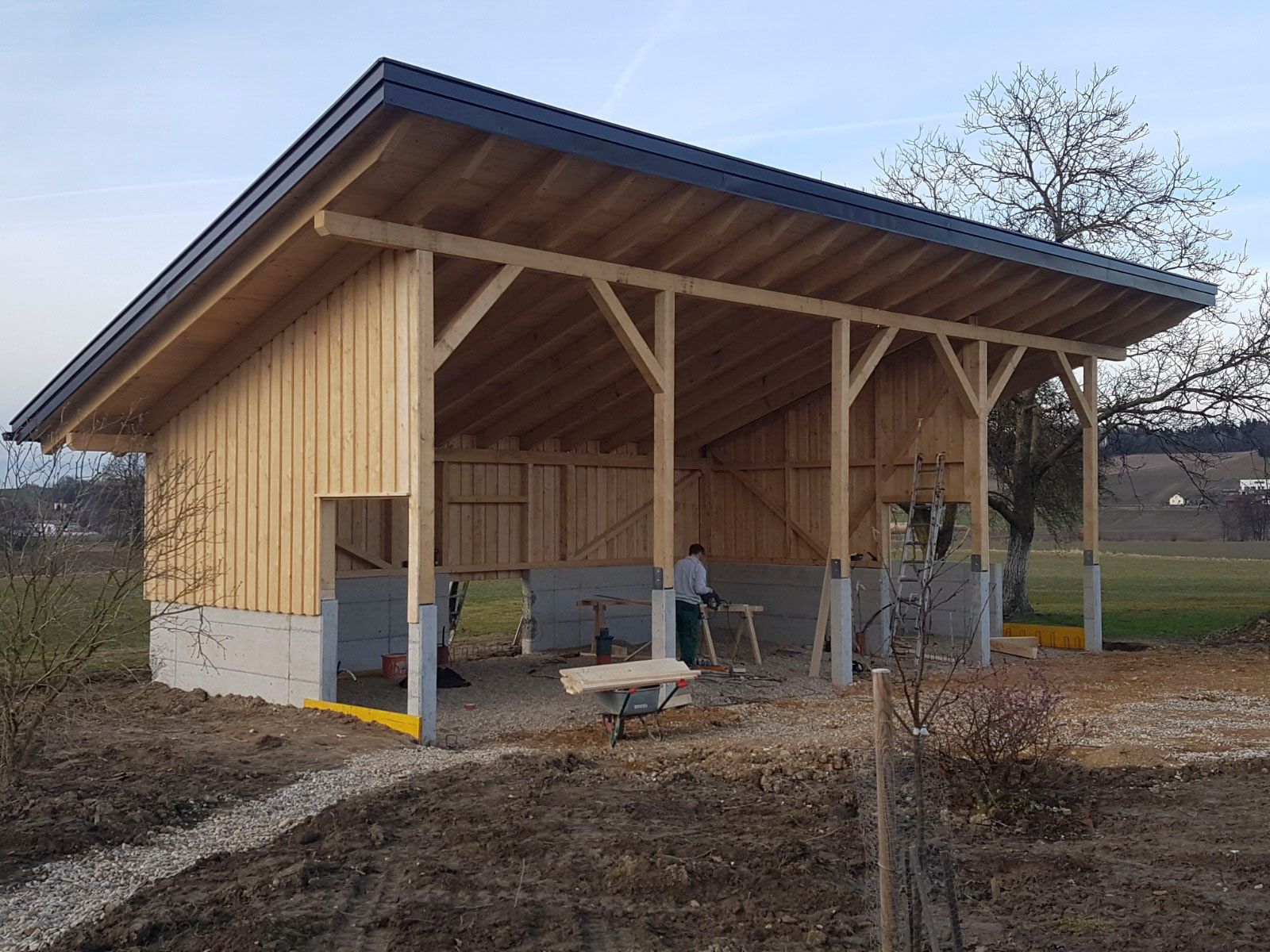 Im Bau befindliche Holzhütte. Helles Holz, schwarzes Dach, Arbeiter im Inneren. Feldumgebung, Kiesweg.