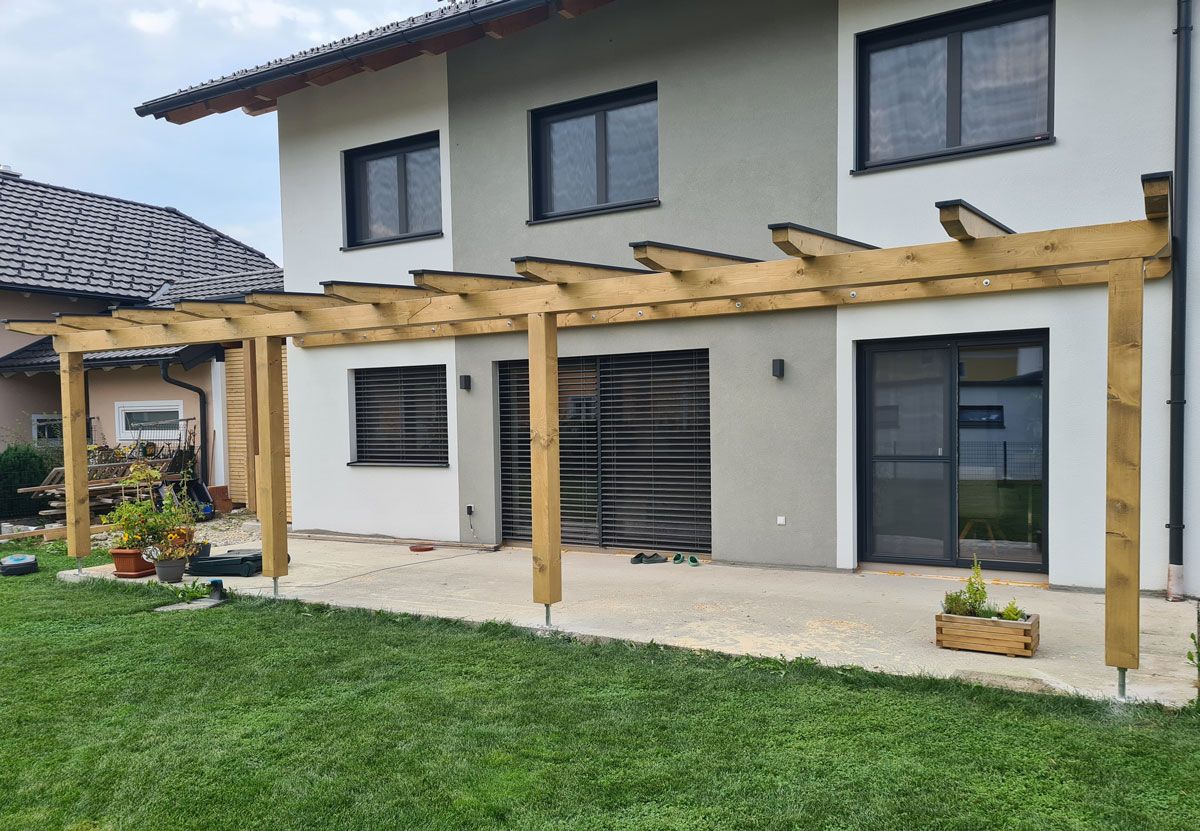 Eine hölzerne Pergola ist an ein zweistöckiges Haus mit Steinterrasse und grünem Rasen angebaut.