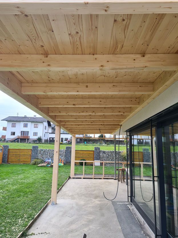 Eine hölzerne Terrassenüberdachung über einer Betonplatte, getragen von Balken und Pfosten, neben einem Haus mit Hof.