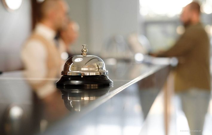 Hotelrezeption mit einer Klingel im Fokus; das Personal ist im Hintergrund unscharf.