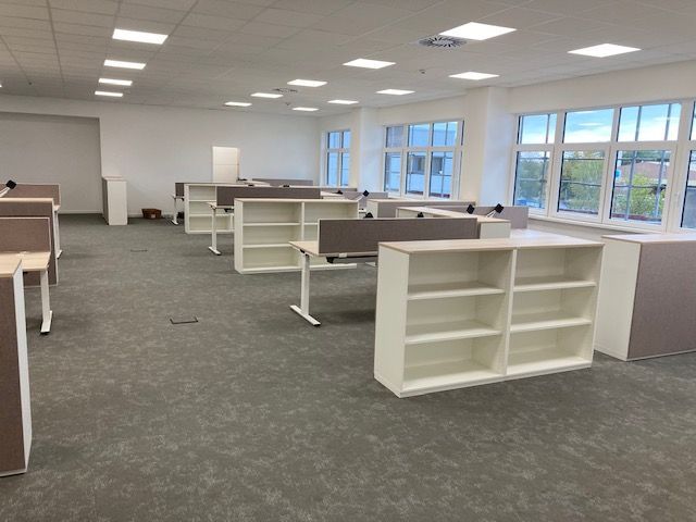 Leerer Büroraum mit Schreibtischreihen, und Bücherregalen. Grauer Teppich, weiße Wände und Möbel in neutralen Farben.