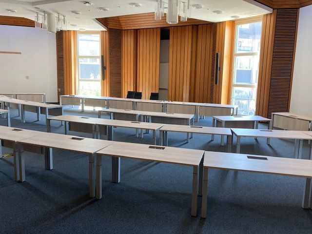 Leerer Konferenzraum mit langen, hellen Tischen, Fenstern und Holzvertäfelung.