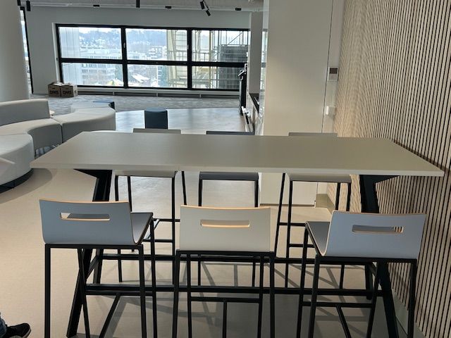 Langer rechteckiger Tisch mit Barhockern in einem modernen Büroraum, große Fenster, helle Farben.