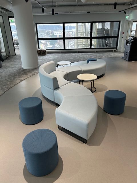 Moderner Bürositzbereich mit geschwungenem, hellblauem Sofa, kleinen runden Tischen und blauen Hockern. Große Fenster bieten einen Blick auf die Stadt.