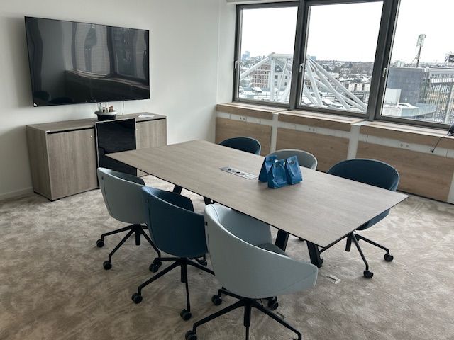 Besprechungsraum mit Tisch, Stühlen, Fernseher, Schrank und Fenstern.