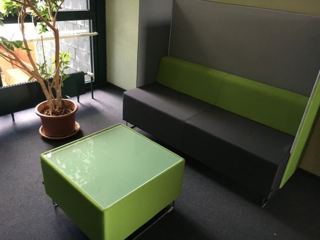 Ein moderner Sitzbereich mit einem grün-grauen Sofa, einem quadratischen Couchtisch und einer Topfpflanze am Fenster.