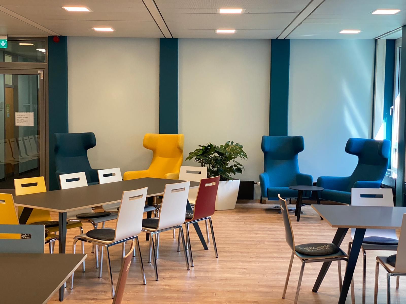 Eine moderne Lounge mit bunten Stühlen, Tischen und einer Pflanze in einem gut beleuchteten Raum.