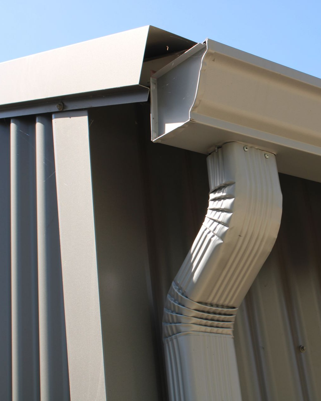 Gouttière et descente pluviale beiges sur un bâtiment en tôle ondulée.