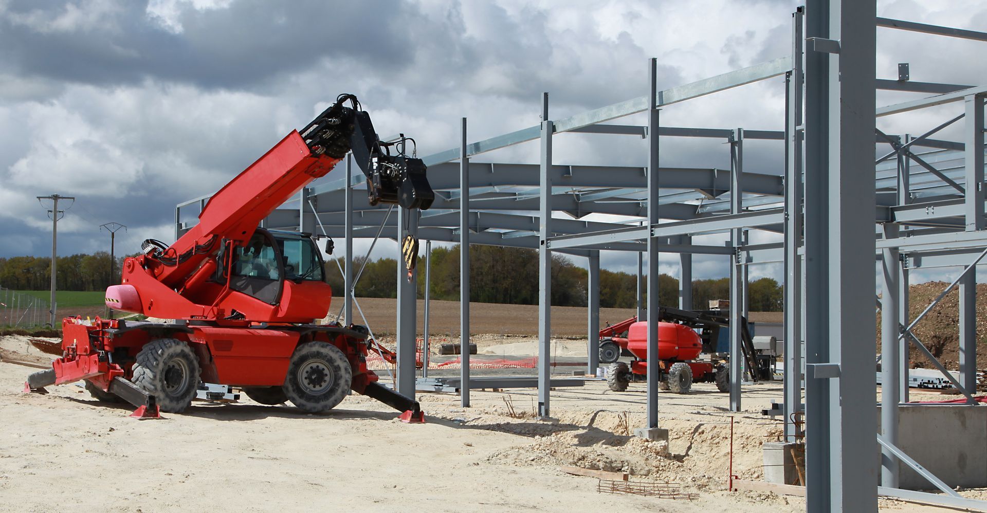 Chariot télescopique rouge soulevant des poutres en acier sur un chantier de construction avec une structure en métal gris.