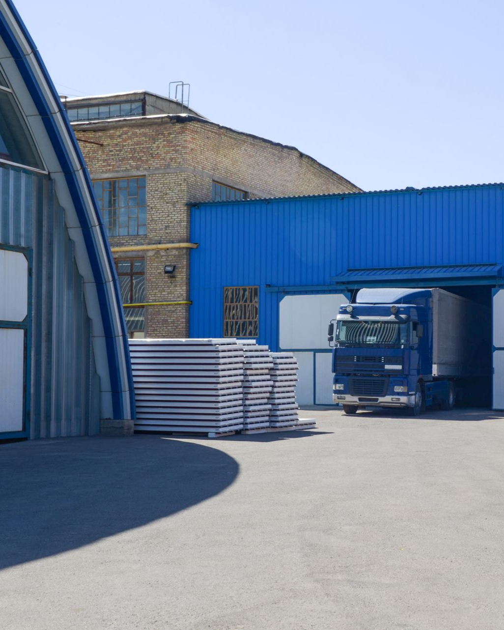 Semi-camion bleu au quai de chargement de l'entrepôt, piles de panneaux sur le sol.