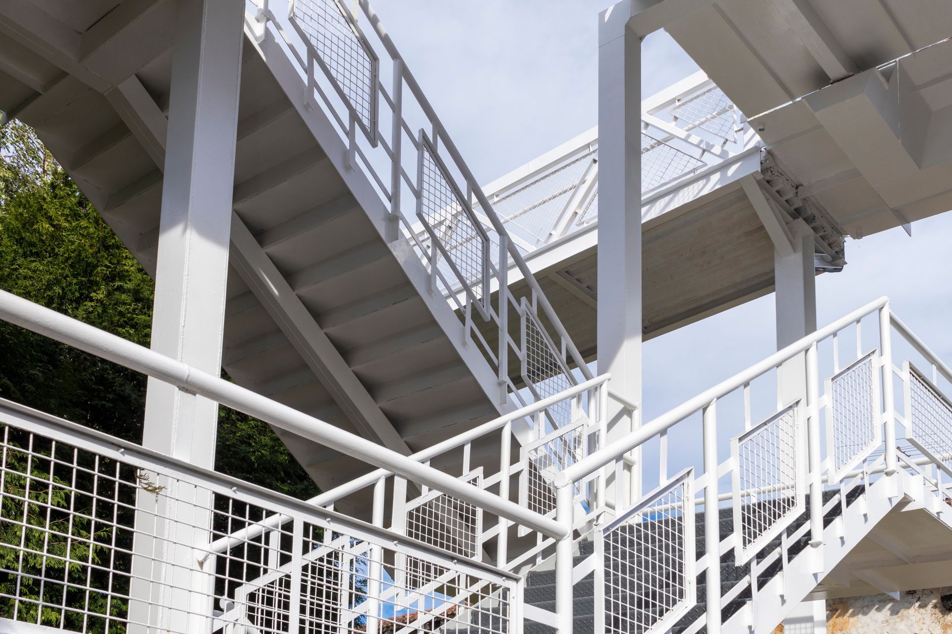 Escalier et passerelle en métal blanc menant à un niveau supérieur contre un ciel nuageux et du feuillage.