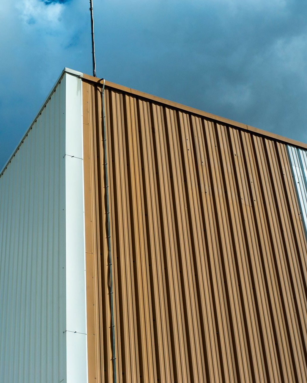 Coin d'un bâtiment commercial avec revêtement en tôle ondulée brune et blanche, un ciel nuageux sombre.