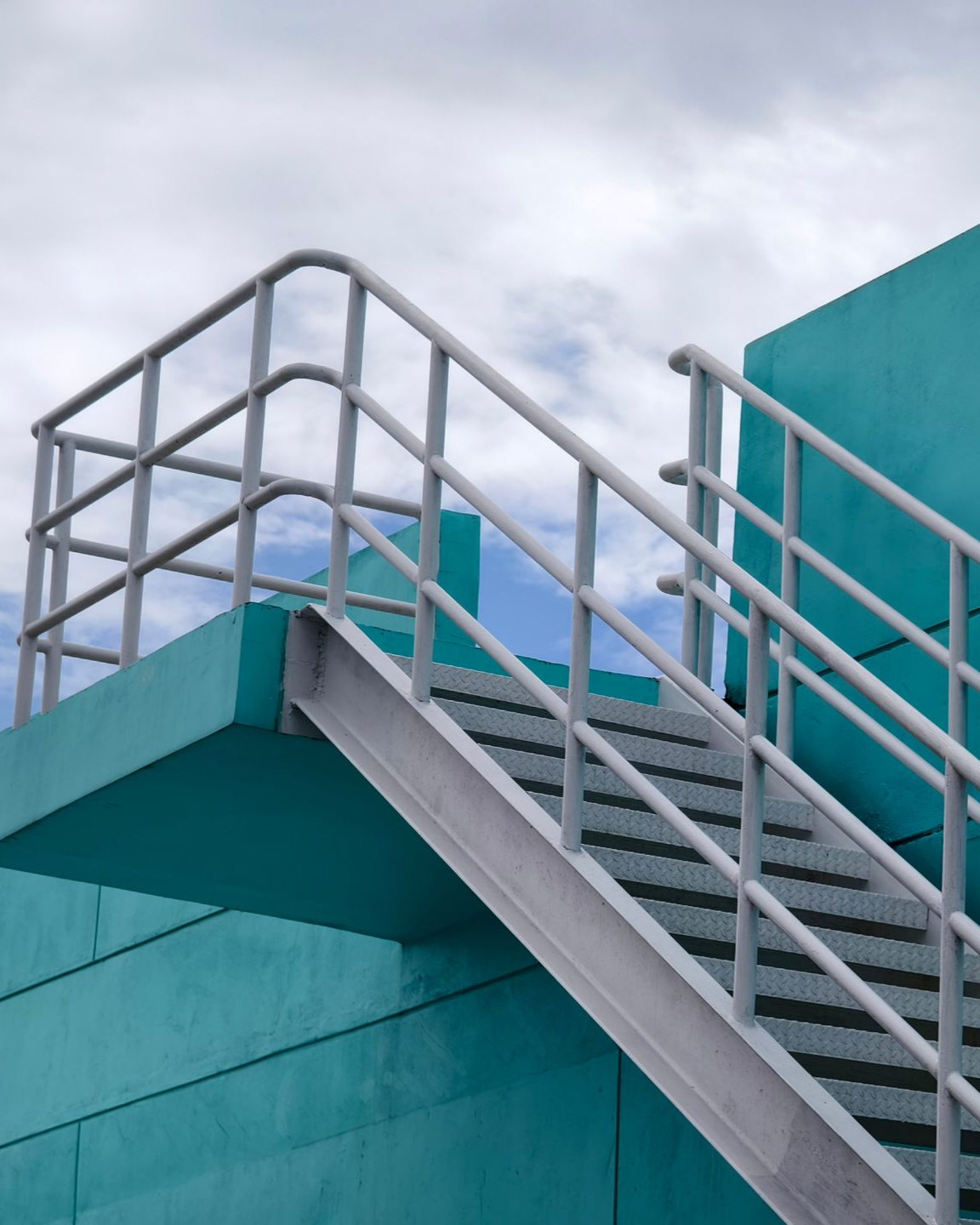 Bâtiment bleu sarcelle avec escalier en métal gris et rampe contre un ciel nuageux.