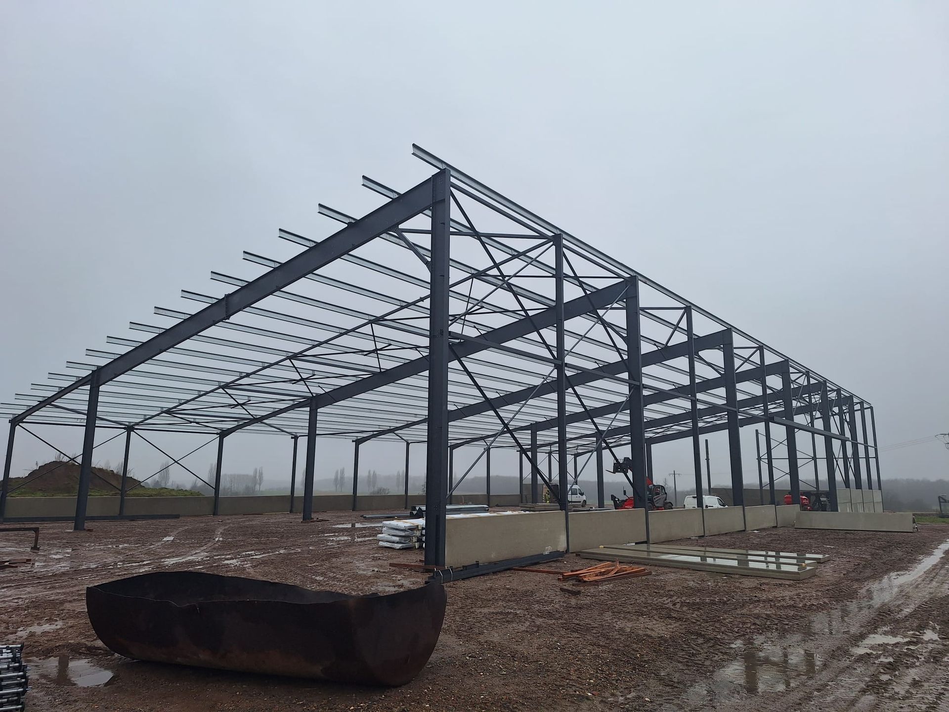 Charpente métallique d'un grand bâtiment en construction sur un terrain en gravier, ciel couvert.