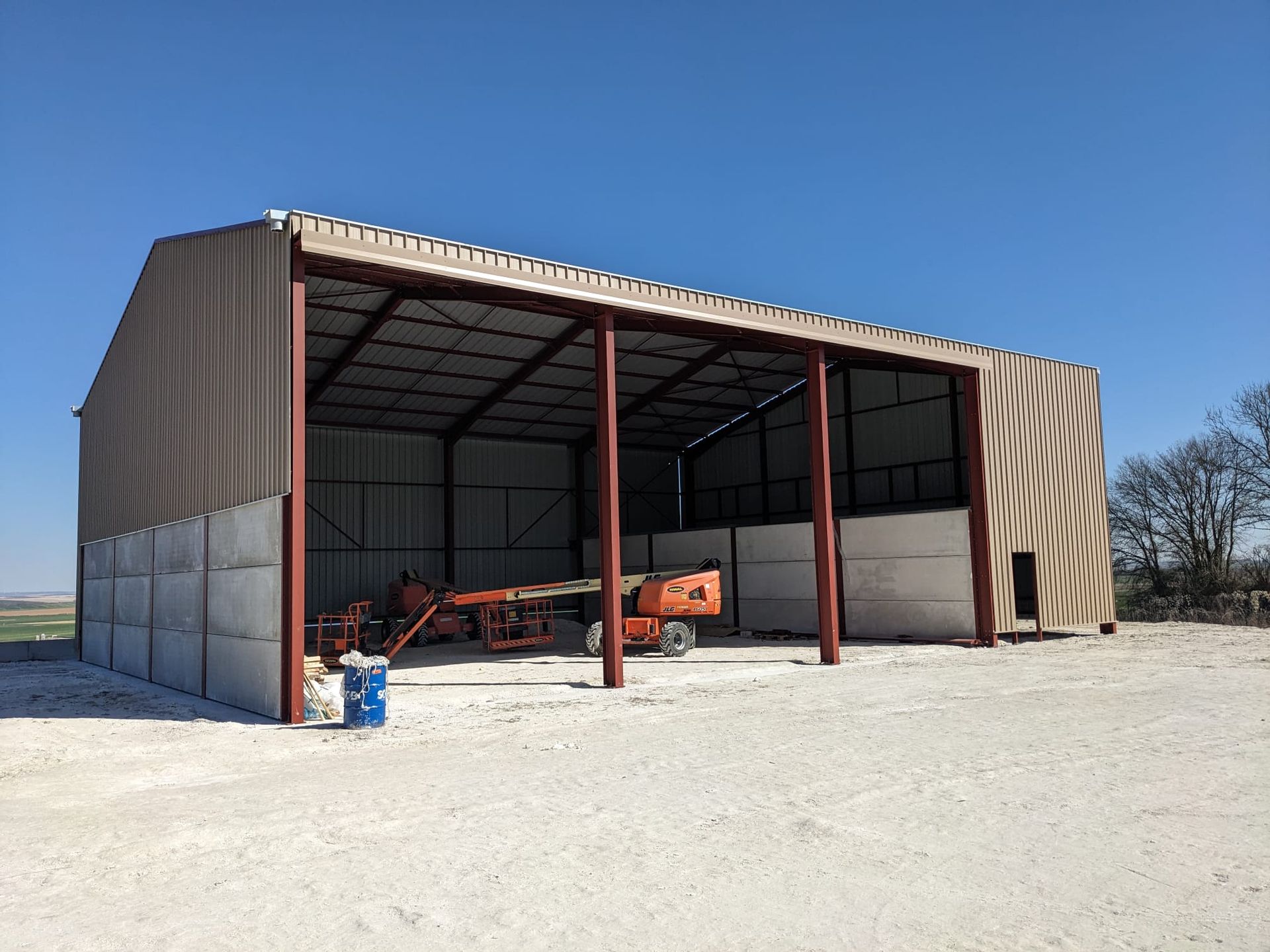 Structure métallique de grange ouverte sur les côtés ; équipement à l'intérieur sur un sol en gravier. Couleurs marron et rouge. Ciel bleu clair.