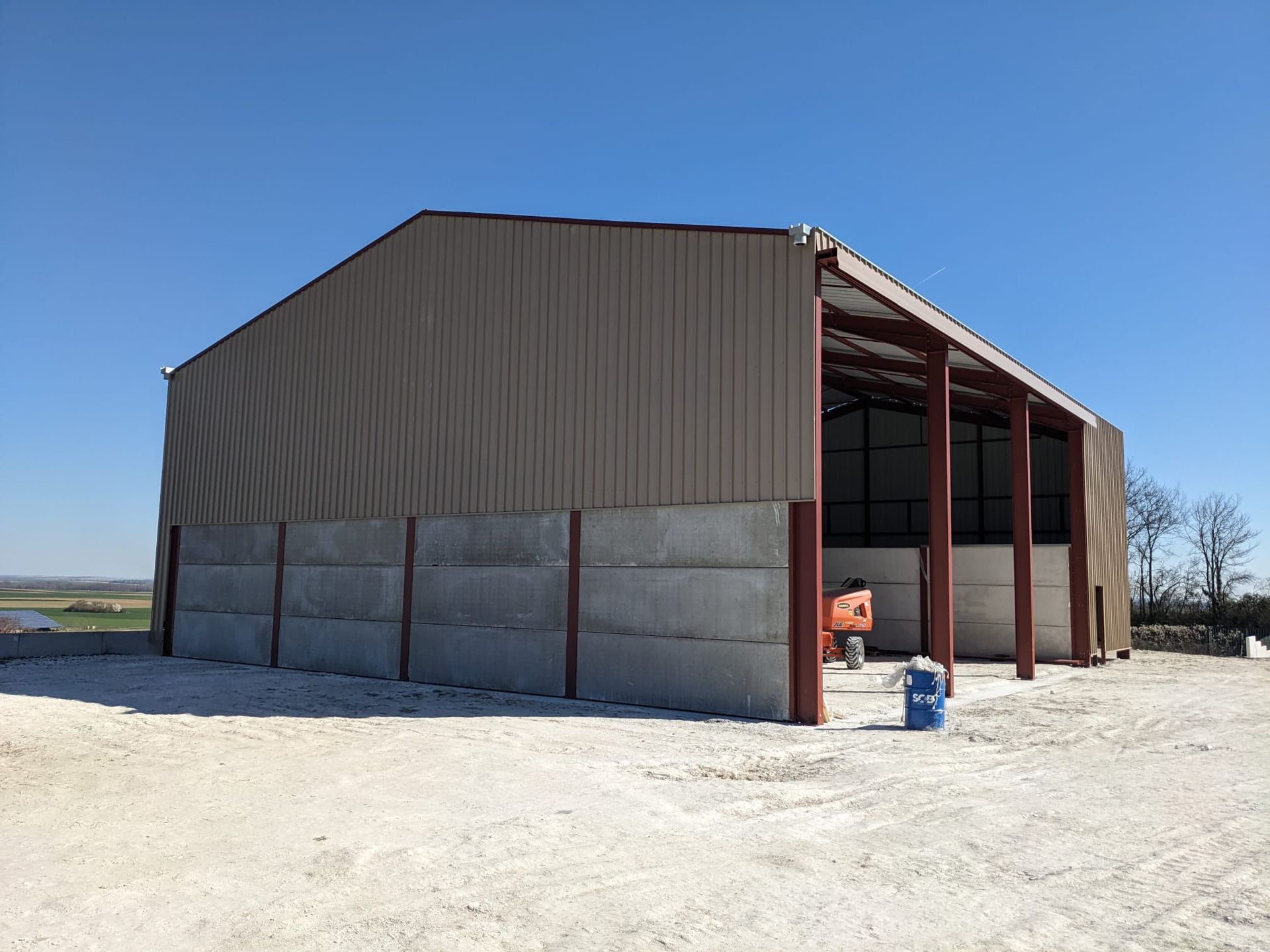 Grange en métal beige avec cadre rouge, mur en béton et côté ouvert par une journée ensoleillée.