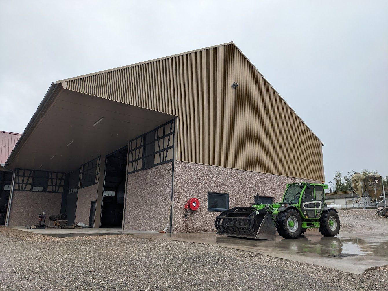 Un tracteur vert garé près d'un entrepôt avec un revêtement en tôle ondulée marron par une journée nuageuse.