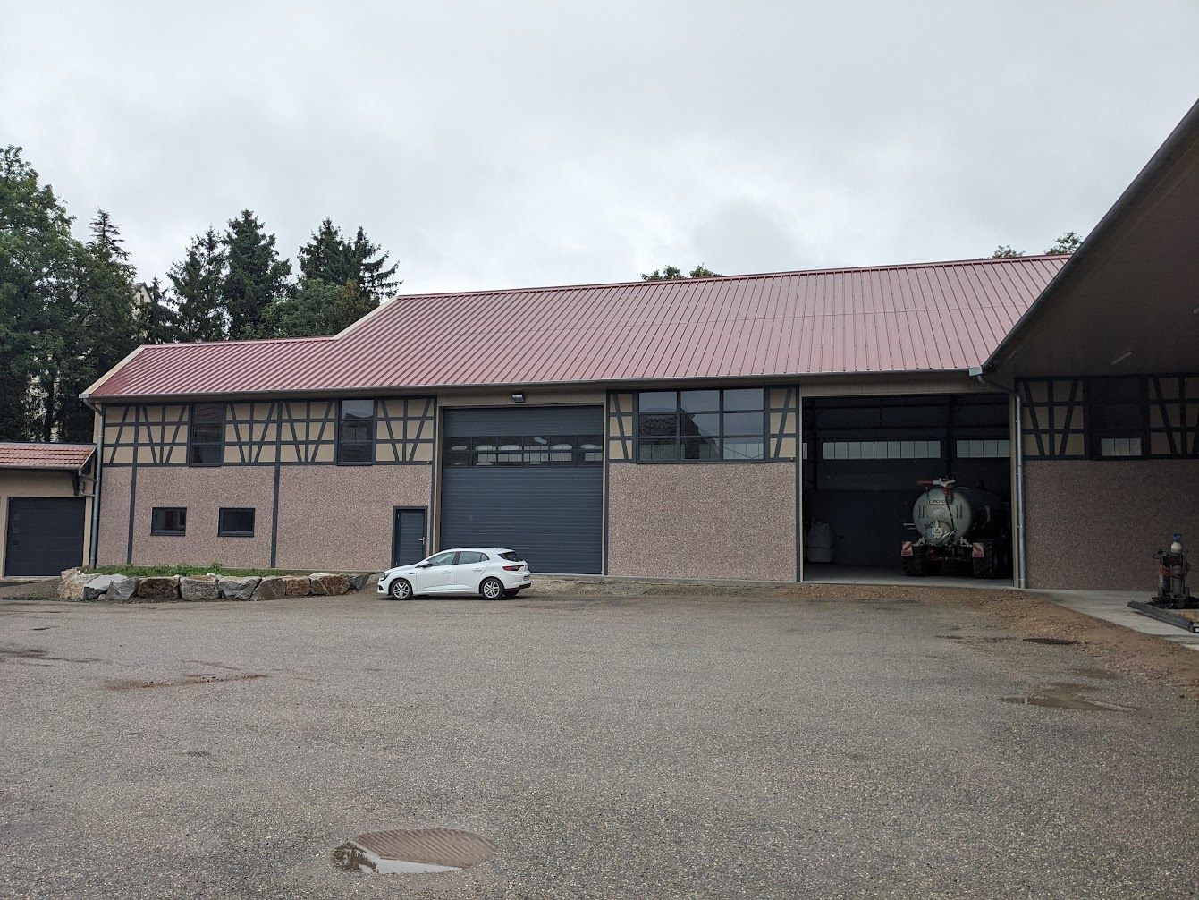 Grange au toit de tuiles rouges, avec deux portes de garage et une petite voiture blanche garée à l'extérieur. Ciel gris et couvert.