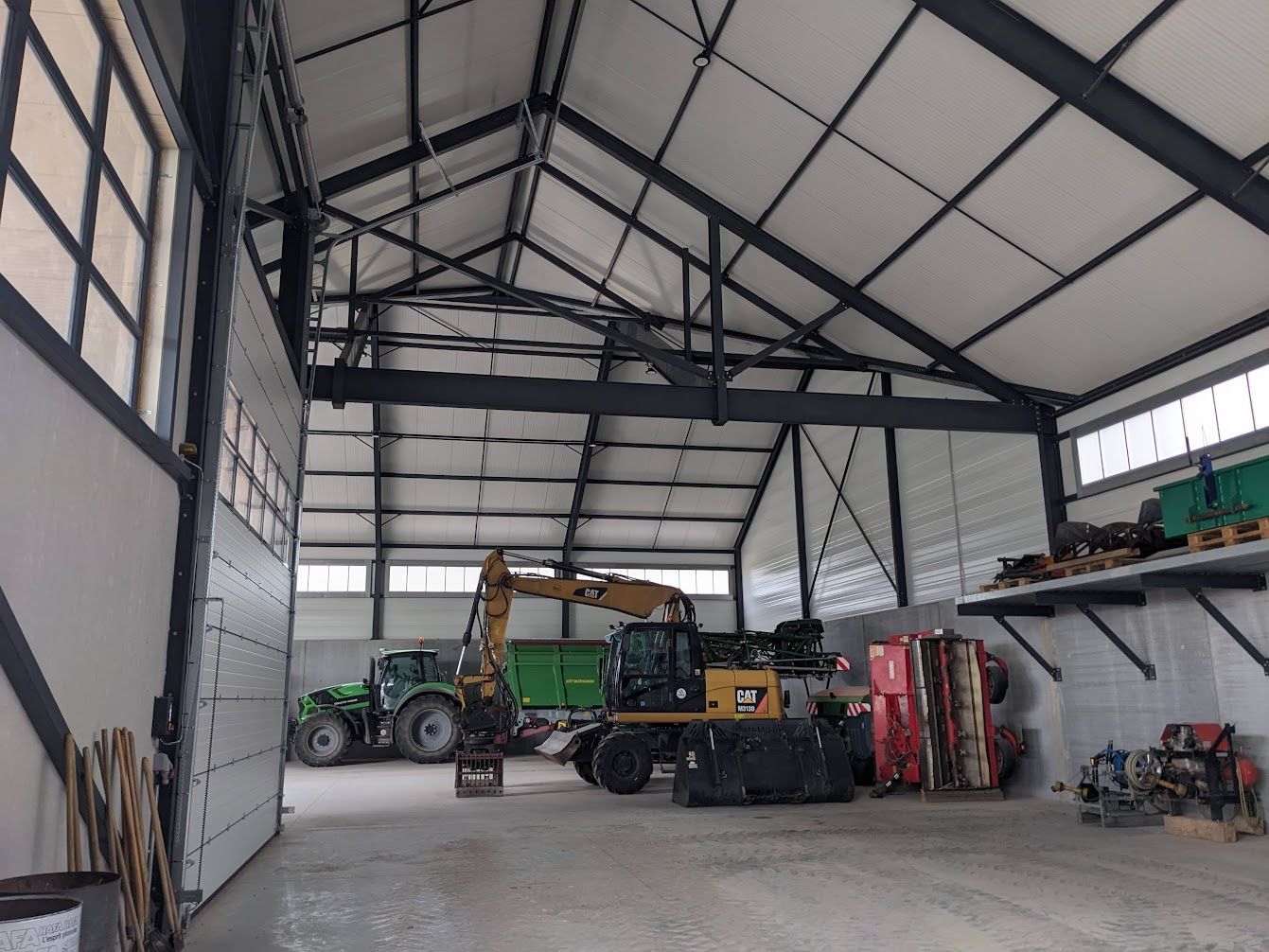 À l'intérieur d'un grand bâtiment métallique, des tracteurs et des machines sont stockés ; une porte de garage est ouverte.
