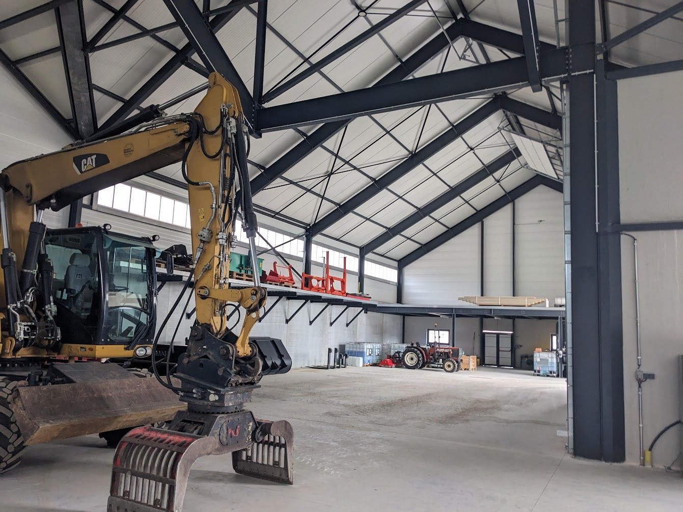 Intérieur d'un grand entrepôt avec une excavatrice jaune. Poutres en acier noires et sol en béton.