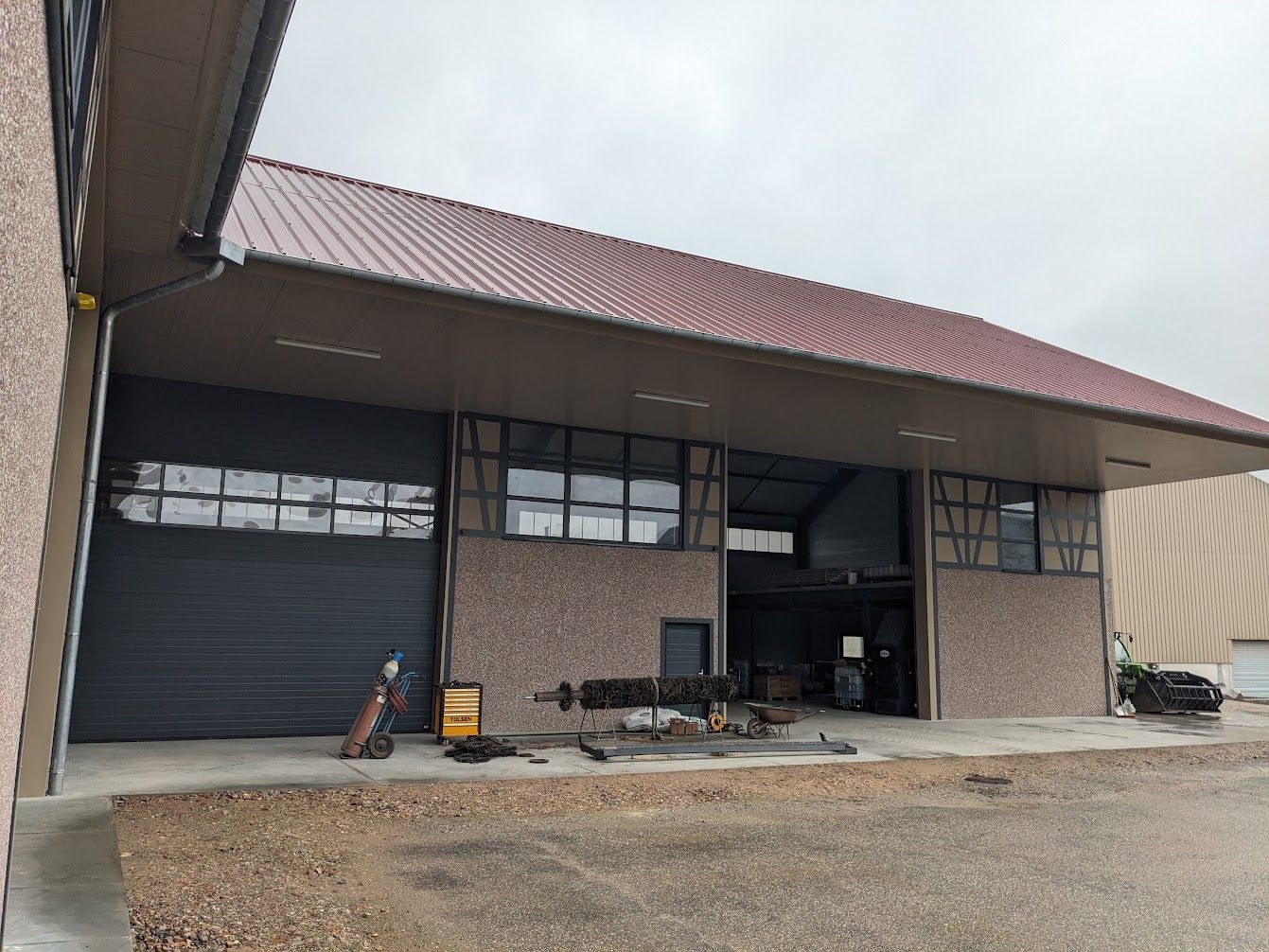 Grand bâtiment avec toit en tuiles rouges et portes de garage. Façade en briques avec outils et équipement à l'avant.