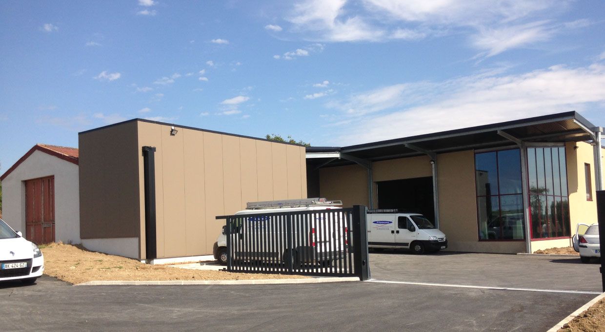 Bâtiment marron avec un portail sombre, une camionnette blanche et un mur de verre sous un ciel partiellement nuageux.