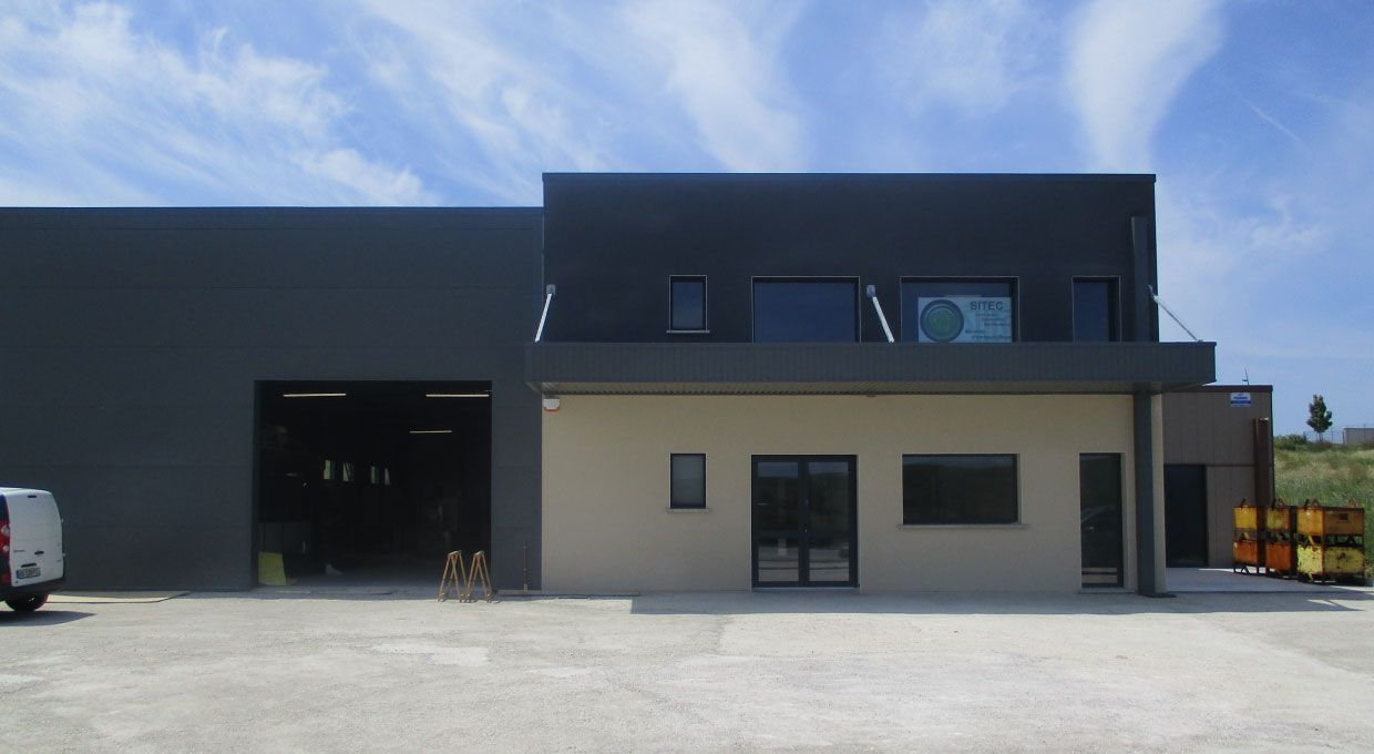 Bâtiment industriel moderne avec façade grise et beige, porte de garage ouverte, camionnette garée, journée ensoleillée.