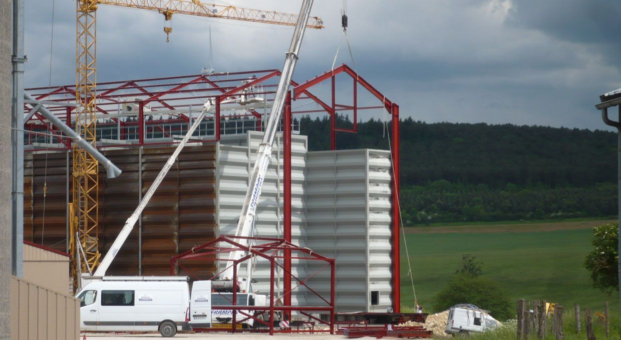 Chantier : une grue soulève une structure en acier rouge sur une grande structure grise. Un camion blanc est garé à proximité.