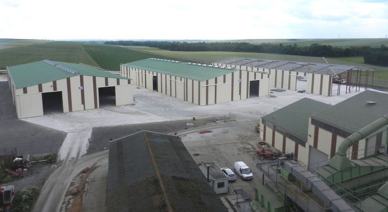 Plusieurs bâtiments industriels aux toits verts, situés sur un terrain en béton, avec des champs en arrière-plan.