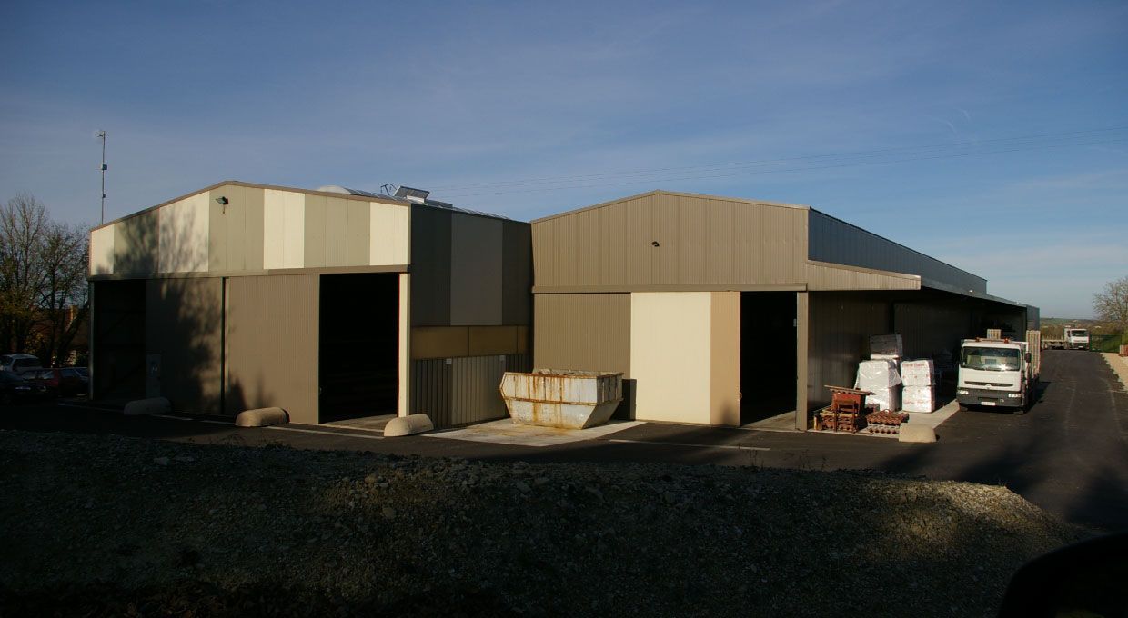 Entrepôt avec portes à baie ouverte; extérieur beige et gris, allée asphaltée, journée ensoleillée.