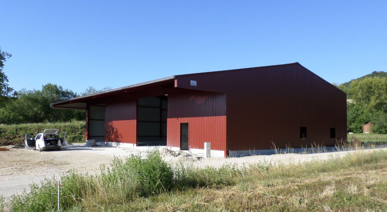 Bâtiment industriel en métal rouge avec entrée ouverte, fenêtres et petite porte sur un terrain en gravier sous un ciel bleu.