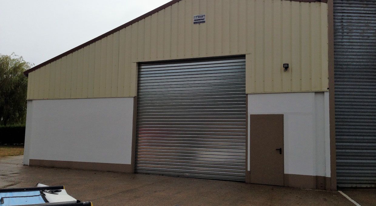 Bâtiment avec porte métallique fermée et petite porte à droite. Extérieur beige et blanc.